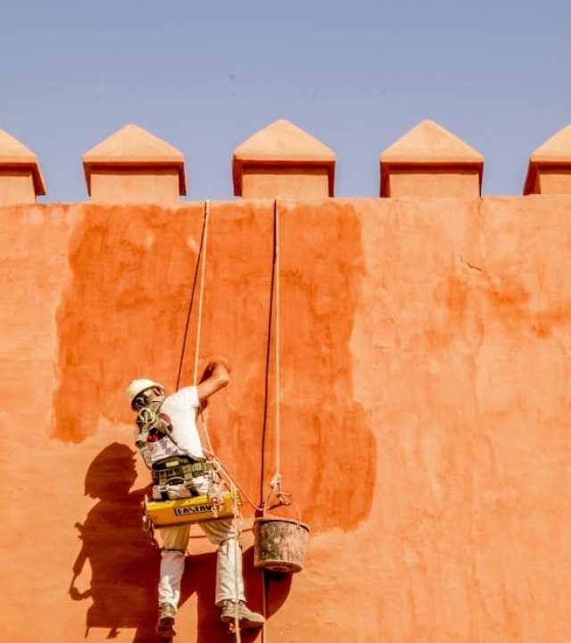 A worker repaints an orange brick wall in the sun, using ropes for support.
